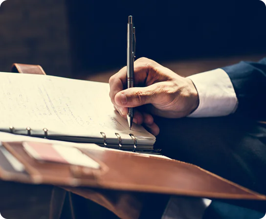 “Close-up of a person in a suit writing with a pen in a ring-bound notebook on their lap, with a leather folder partially visible in the foreground.”